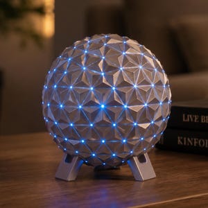 May include: A spherical lamp with a silver geodesic surface featuring a repeating triangular pattern. Small, bright blue LED lights are embedded at the intersection points of the geometric structure. The lamp is supported by three short, sturdy legs.