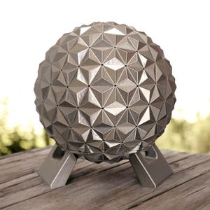 Op de afbeelding: Een zilverkleurig bolvormig object met een geometrisch patroon van onderling verbonden driehoeken over het oppervlak. De bol wordt ondersteund door een stevige, bijpassende zilverkleurige basis met drie afzonderlijke poten.