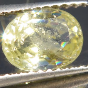 May include: An oval-cut, pale yellow gemstone held by metal tweezers. The stone features numerous facets that reflect light, creating bright highlights and internal prismatic colour refractions throughout its translucent, crystalline structure.