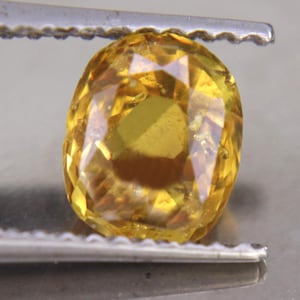 May include: A cushion-cut yellow gemstone with a faceted surface that reflects light. The stone displays a vibrant golden hue and is held by a pair of metal tweezers, showcasing its transparent clarity and intricate geometric cut pattern.