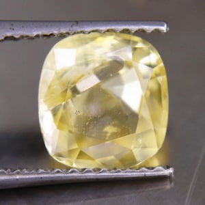May include: A cushion-cut yellow gemstone with a faceted surface held by metal tweezers. The stone displays a translucent, pale lemon-yellow hue with internal light reflections and sharp geometric edges across its polished top surface.