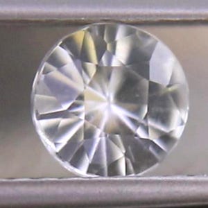 May include: A round, clear gemstone with numerous facets, reflecting light. The gemstone is held between two metal bars, showcasing its brilliance and cut. The gem appears to be colorless, with sharp angles and a polished finish.