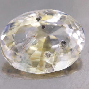 May include: A faceted, oval-shaped gemstone with a pale yellow hue. The gem has internal inclusions and a brilliant cut, reflecting light. The stone is set against a neutral background, highlighting its clarity and color. This gemstone is suitable for jewelry.