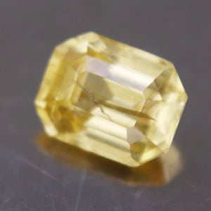 May include: A faceted yellow gemstone with an emerald cut, displaying a translucent appearance and a rectangular shape with beveled edges. The stone exhibits a bright, pale yellow hue and internal light reflections across its polished surfaces.
