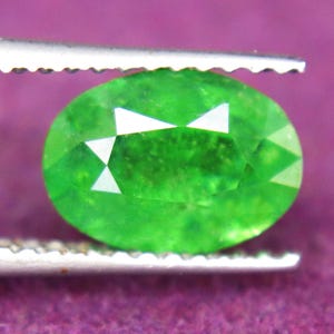 May include: An oval-cut, vibrant green gemstone held by silver-toned metal tweezers. The stone features a faceted surface that reflects light, showcasing a bright, saturated emerald-like hue with internal clarity and distinct geometric facets.