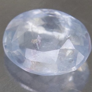 Könnte beinhalten: Ein facettierter ovaler Edelstein in einer blassen, durchscheinenden hellblauen Farbe. Der Stein weist mehrere geometrisch geschliffene Oberflächen auf, die das Licht reflektieren und ein klares, kristallines Aussehen mit subtilen Einschlüssen erzeugen.