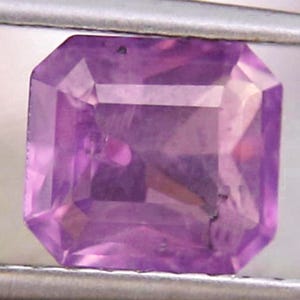 May include: A faceted, rectangular-cut gemstone in shades of purple. The gem is held by a metal tool, showcasing its geometric facets and internal clarity. The gem's color ranges from light to deep purple.