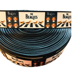 May include: A roll of black ribbon with a repeating pattern featuring the text "The Beagles" and illustrations of beagle dogs. The design includes dogs crossing a crosswalk and beagle faces with sunglasses.