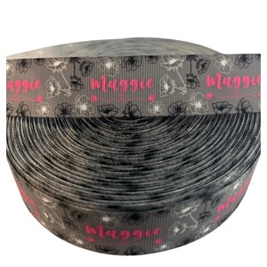 May include: A roll of gray ribbon with a floral pattern and the name "Maggie" in pink. The ribbon features a repeating design of white and black flowers. The spool is tightly wound, showcasing the ribbon's texture and design.