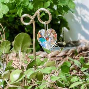 Puede incluir: Un prisma de cristal en forma de corazón de metal dorado que cuelga de un gancho de metal dorado. El prisma es iridiscente y refleja la luz.