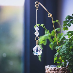 May include: A gold metal plant hanger with a crystal prism hanging from it. The prism is iridescent and has a teardrop shape. The hanger is attached to a potted plant with green leaves.