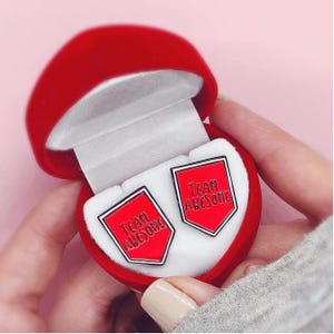 May include: A pair of red and black enamel cufflinks with the text "TEAM AWESOME" in white, presented in a red heart-shaped jewelry box with a white interior. The cufflinks are shield-shaped.