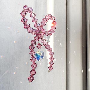 May include: A pink crystal bow with a small crystal heart hanging from the bottom. The bow is made of many small crystals strung together on a wire.
