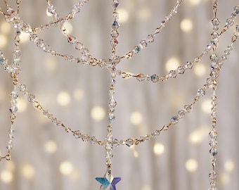 May include: A crystal garland with iridescent star accents hanging against a blurred background of white lights. The garland is made of small, clear crystals strung on a gold wire.
