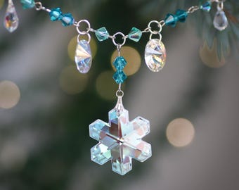 May include: A crystal snowflake ornament with teal and clear crystal beads hanging from a silver chain. The ornament is hanging from a Christmas tree.
