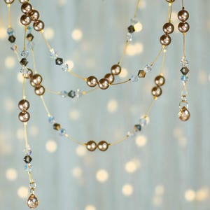 May include: A double strand of brown pearl and crystal beads on a gold wire. The beads are hanging from a branch against a blurred background of white lights.