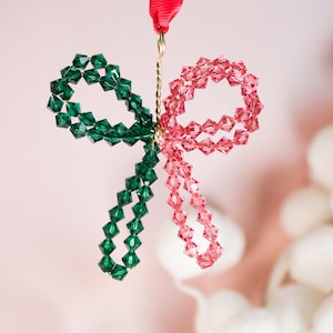 May include: A Christmas bow-shaped ornament made from green and pink faceted beads. The ornament is suspended by a red ribbon and a gold-coloured wire, creating a festive decoration.