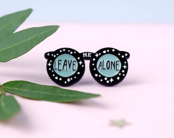 May include: A black enamel pin in the shape of sunglasses with the words "LEAVE ME ALONE" in white lettering on a teal background. The pin has white star and dot accents. The background is a soft pink with green leaves.