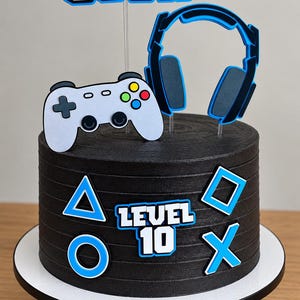 Op de afbeelding: Een zwarte taart versierd met blauwe en witte gaming-toppers. De taart bevat de tekst "LEVEL 10" en symbolen zoals een driehoek, cirkel, vierkant en X. De toppers bestaan uit een witte gamecontroller, een blauwe koptelefoon en de naam "JOSH".