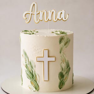 Può includere: Una torta cilindrica con glassa color bianco sporco, decorata con motivi a foglie verdi dipinte e una croce bianca centrale con bordo dorato. Un topper per torta in corsivo bianco con bordi dorati mostra il nome Anna in cima.