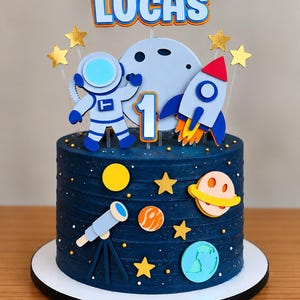 Könnte beinhalten: Ein dunkelblauer Kuchen mit Weltraum-Dekorationen wie Planeten, einem Teleskop und Sternen. Ein Tortenstecker zeigt den Namen LUCAS in blauen Buchstaben, einen Astronauten, einen Mond, eine Rakete und die Zahl 1 auf durchsichtigen Stäben.