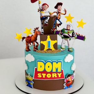 May include: A round cake decorated with light blue and brown frosting, featuring Toy Story character cutouts including Woody, Jessie, Buzz Lightyear, Bullseye, and Mr. Potato Head. The cake displays the text DOM STORY and a yellow star with the number 1.