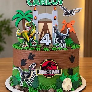 Könnte beinhalten: Ein runder Kuchen mit braunem und grünem Zuckerguss, verziert mit Dinosaurier-Motiven wie einem T-Rex, Raptoren, einem Vulkan, einer Palme, einem Tor, dem Jurassic Park-Logo und einem Aufsteller mit dem Namen CARLOS und der Zahl 4.