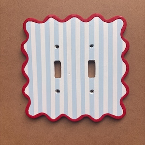Puede incluir: Una placa de pared para interruptor doble con un borde festoneado pintado en rojo brillante. La superficie central muestra un patrón repetitivo de rayas verticales de color azul claro y blanco. La placa incluye dos aberturas rectangulares.