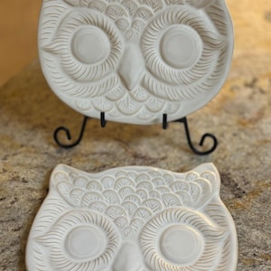 May include: Two off-white owl-shaped decorative plates. The plates have embossed details, including the eyes, feathers, and beak. One plate is displayed on a black metal stand. The plates are likely for decorative purposes.