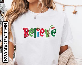 May include: White t-shirt with the word "Believe" in colorful, festive lettering. The shirt is paired with red and white plaid pajama pants. A mug with a Santa Claus design is held in the hand. The background includes a decorated Christmas tree.
