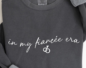 May include: Dark gray sweatshirt with the text "in my fiancée era" in white script, along with two interlocking rings. The sleeves have gray, fuzzy cuffs. The sweatshirt is a comfortable, casual style.