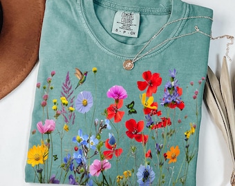 May include: A sage green crewneck t-shirt featuring a vibrant graphic print of various wildflowers, a hummingbird, and a butterfly. The shirt has a Comfort Colors brand label at the inner neckline and is displayed flat with a delicate silver necklace.