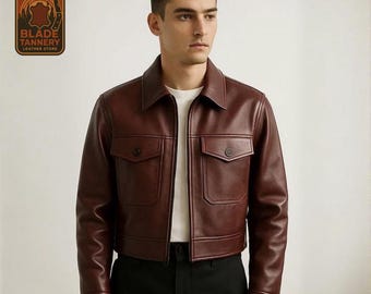 May include: A burgundy leather jacket with two front pockets and a collared neckline. The jacket has a button closure and is worn over a white shirt. The logo for Blade Tannery Leather Store is in the upper left corner.