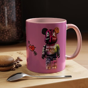 Könnte beinhalten: Eine rosa Keramiktasse mit hellrosa Henkel und Innenraum. Die Tasse zeigt eine farbenfrohe Grafik einer bärenartigen Figur mit den Worten "NO RISK NO NAMA" und einer Lippe mit einem Pfeil. Ein Löffel und ein Macaron sind im Vordergrund.