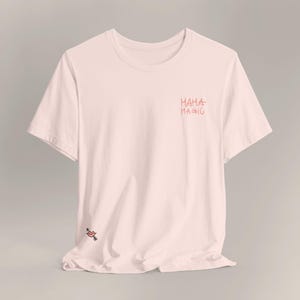 Könnte beinhalten: Hellrosa T-Shirt mit dem Schriftzug "Mama Magic" in roter Schrift auf der Brust. Eine kleine rote Lippen-Grafik mit Pfeil befindet sich unten links. Das Shirt hat einen Rundhalsausschnitt und kurze Ärmel.