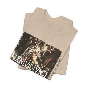 Könnte beinhalten: Ein beiges T-Shirt mit einem dunklen, malerischen Grafikdruck. Das Design zeigt eine stilisierte Figur, rote Blattakzente und den Text "VENI.VIDI.VICI" in fetten weißen Buchstaben, ergänzt durch den Schriftzug "esto por la gloria" darunter.