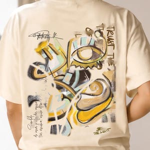 Könnte beinhalten: Cremefarbenes T-Shirt mit einem abstrakten Design in Gelb, Schwarz und Blau. Auf der Rückseite des Shirts stehen die Worte "TRUST THE" und "BE THE LIGHT" sowie eine Unterschrift und weiterer Text.