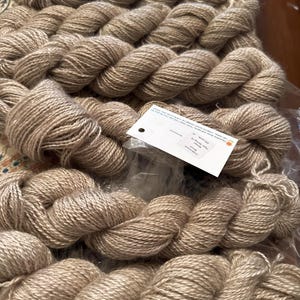 May include: Close-up of several skeins of light brown yarn. The yarn is tightly wound and has a slightly textured appearance. A white tag is visible, possibly indicating the yarn's details. The yarn is likely for knitting or crocheting projects.