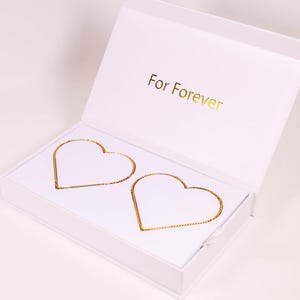 Puede incluir: Una caja de regalo blanca con las palabras "For Forever" en letras doradas. En el interior, dos contornos dorados en forma de corazón se muestran sobre un fondo blanco, lo que sugiere una joya o recuerdo.