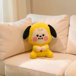 以下が含まれることがあります： 黒い耳、笑顔の顔、ピンクの頬を持つ漫画のキャラクターの形をした黄色のぬいぐるみ。 おもちゃは手を合わせており、胸に赤いボタンが付いています。 白いソファに座っています。