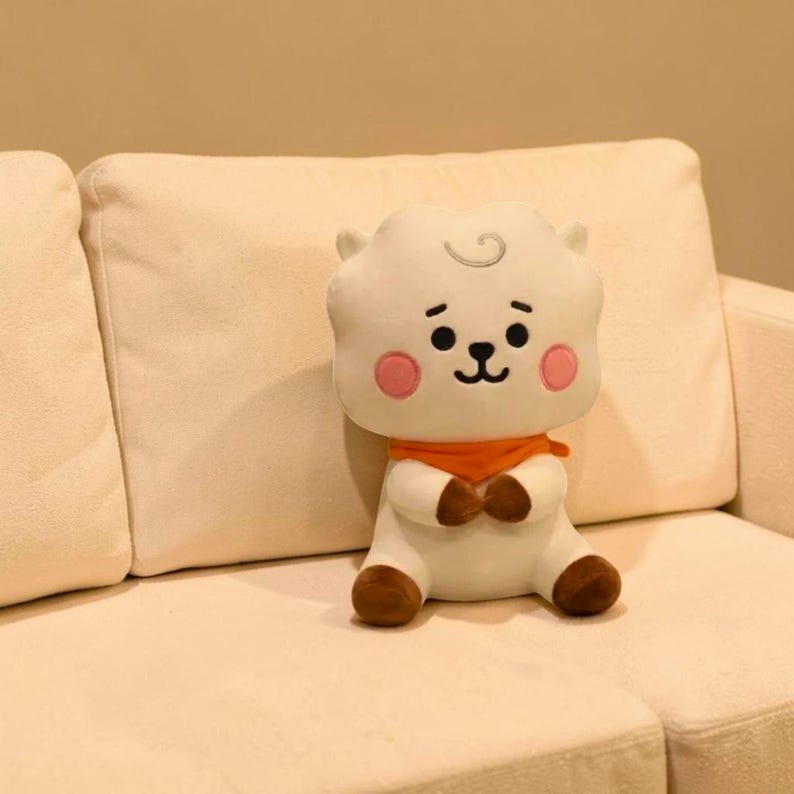以下が含まれることがあります： 白い雲のようなキャラクターの形をしたぬいぐるみ。笑顔の顔、ピンクの頬、茶色の足と手があります。オレンジ色のスカーフを着て、クリーム色のソファに座っています。