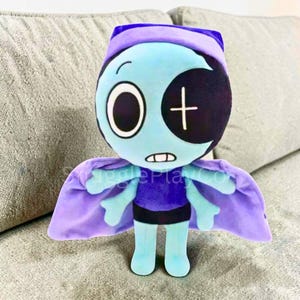 May include: A plush doll with a blue face, large eyes, and a black patch over one eye. It has a purple cape and a matching hat. The doll's body is blue, with a purple shirt and black shorts. The text "SnugglePlayCo" is visible on the cape.