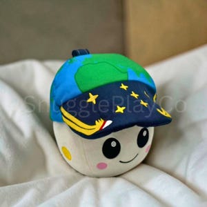 May include: A plush toy shaped like a smiling moon wearing a blue cap with a green earth design and yellow stars. The moon has pink cheeks and a yellow spot. The cap has a yellow and white design on the brim.