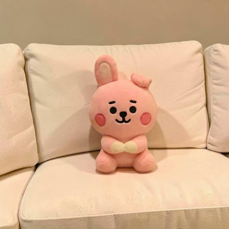 以下が含まれることがあります： 笑顔、ピンクの頬、小さな蝶ネクタイが特徴のピンクのぬいぐるみウサギのおもちゃ。ウサギは片方の耳が上がっていて、片方の耳が垂れ下がっています。おもちゃは白いソファの上に座っています。