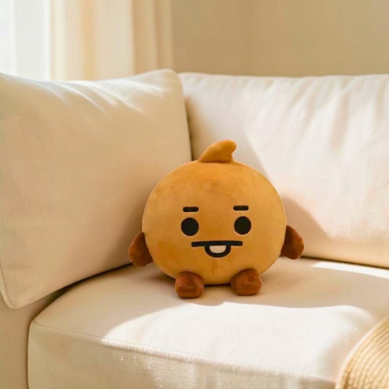以下が含まれることがあります： 丸いオレンジ色のぬいぐるみキャラクターで、上部に小さなヒレのような突起があります。キャラクターは、黒い目、笑顔の口、小さな腕と足を持つシンプルな顔をしています。白いソファの上にあります。