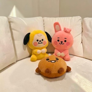 以下が含まれることがあります： 3つのぬいぐるみ。黒い耳と笑顔の顔をした黄色いキャラクター、片方の耳が上がったピンクのウサギ、小さな腕と笑顔の顔をした茶色の丸いキャラクター。おもちゃは白いソファの上にあります。