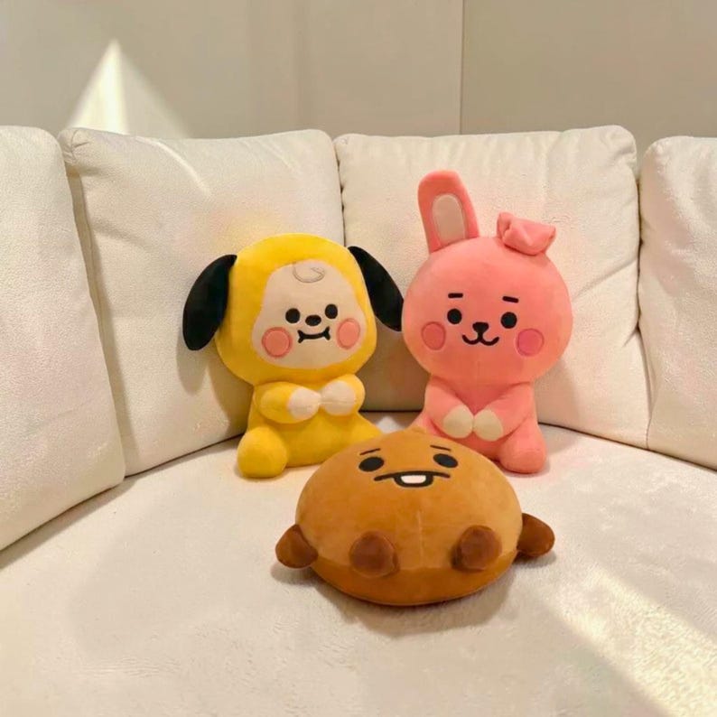 以下が含まれることがあります： 3つのぬいぐるみ。黒い耳と笑顔の顔をした黄色いキャラクター、片方の耳が上がったピンクのウサギ、小さな腕と笑顔の顔をした茶色の丸いキャラクター。おもちゃは白いソファの上にあります。