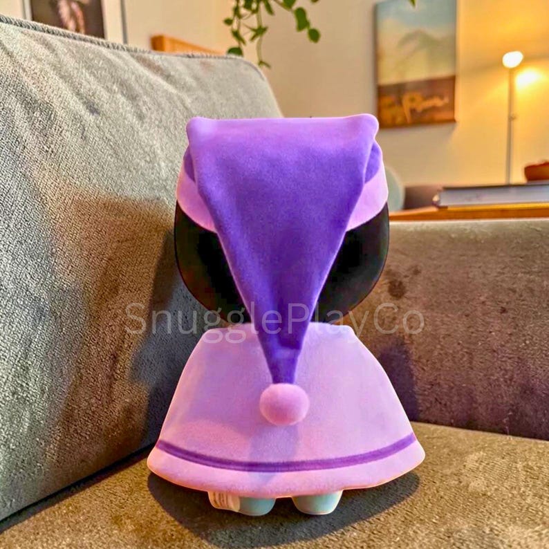 May include: A soft toy character wearing a purple nightcap and a light purple dress. The toy has a black face and small blue feet. The text "SnugglePlayCo" is visible on the front of the toy.