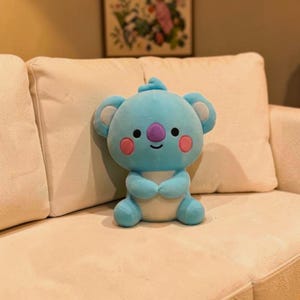 以下が含まれることがあります： 白いお腹、ピンクの頬、紫の鼻を持つ、水色のぬいぐるみコアラ。 コアラは大きくて丸い耳をしており、クリーム色のソファに座っています。 おもちゃは柔らかく、抱きしめたくなるようなものです。