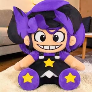 Pode incluir: Uma boneca de pelúcia com um design de desenho animado. A boneca tem cabelo roxo e preto, olhos grandes e um sorriso largo. Ela usa uma roupa preta com detalhes de estrelas amarelas. A boneca está sentada em um tapete branco.